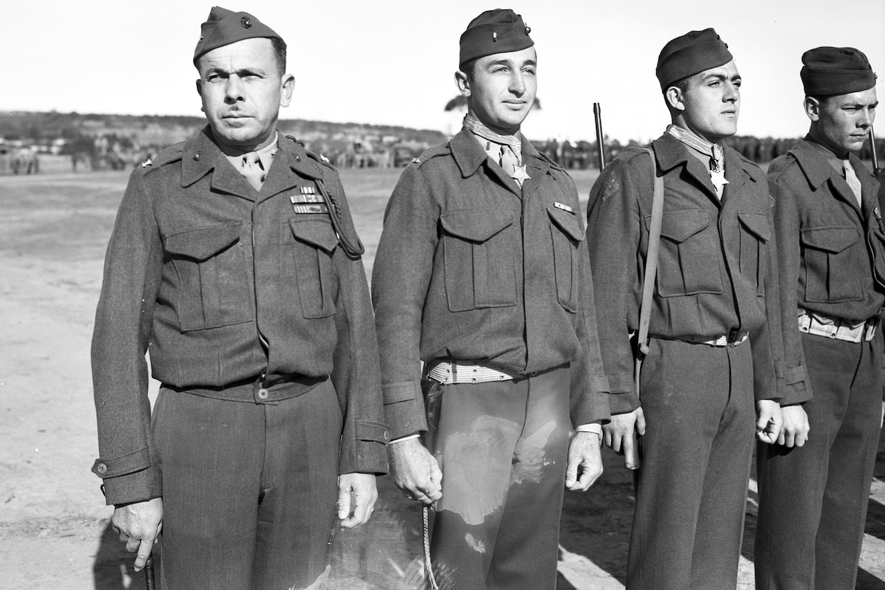 Four Marines stand in a row on what appears to be an airfield. The two men in the middle are wearing Medals of Honor.