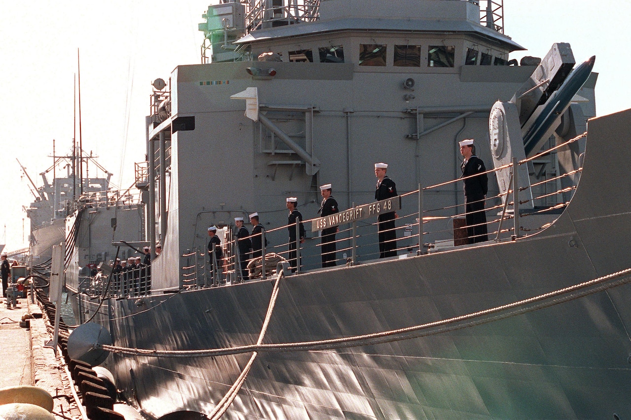 Several sailors in dress blues stand at the rail of a large, docked guided missile frigate. In small letters you can see the ship’s name, USS Vandegrift FFG 48.