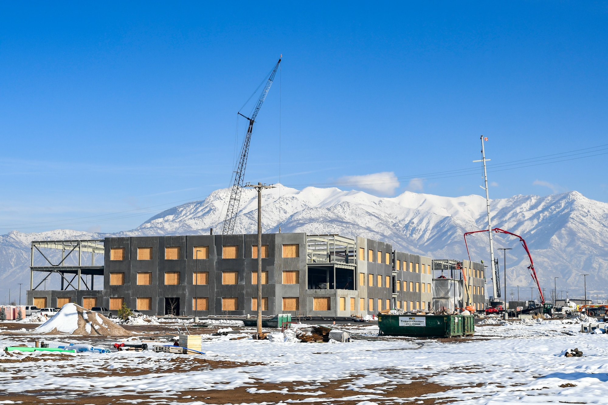The construction site of the Northrup Grumman Roy Innovation Center situated adjacent to the Hill Aerospace Museum appears with snow covered ground, blue skies, and snow-capped mountains in the background.