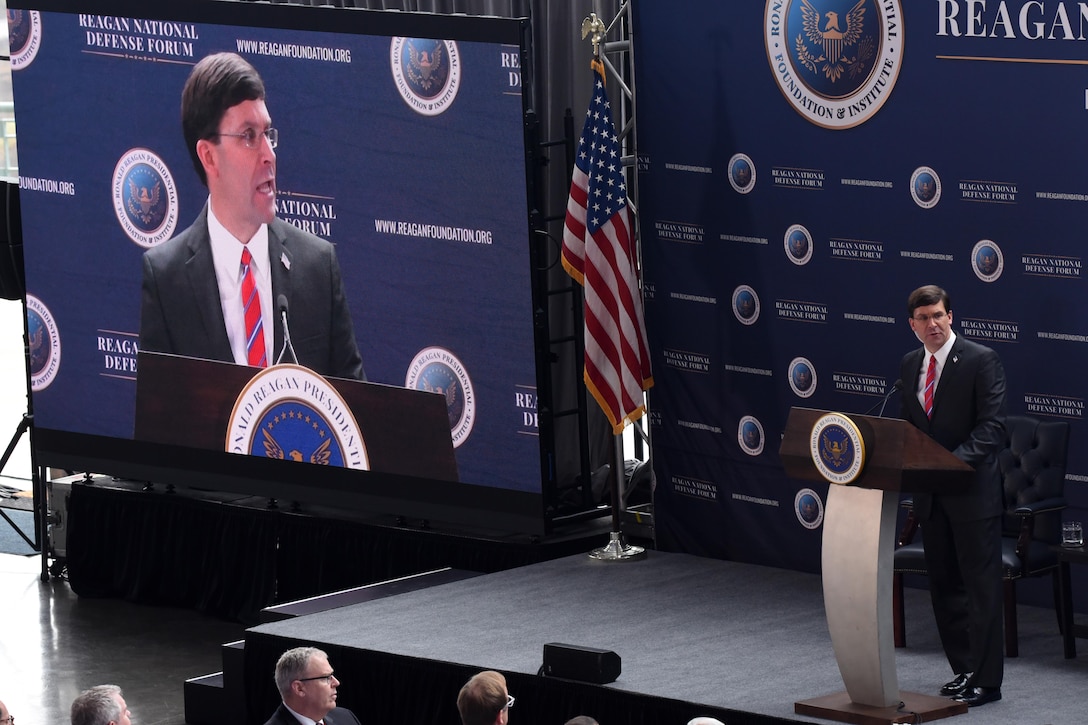 Defense Secretary Dr. Mark T. Esper speaks at a podium while also being shown on a large screen to his right.