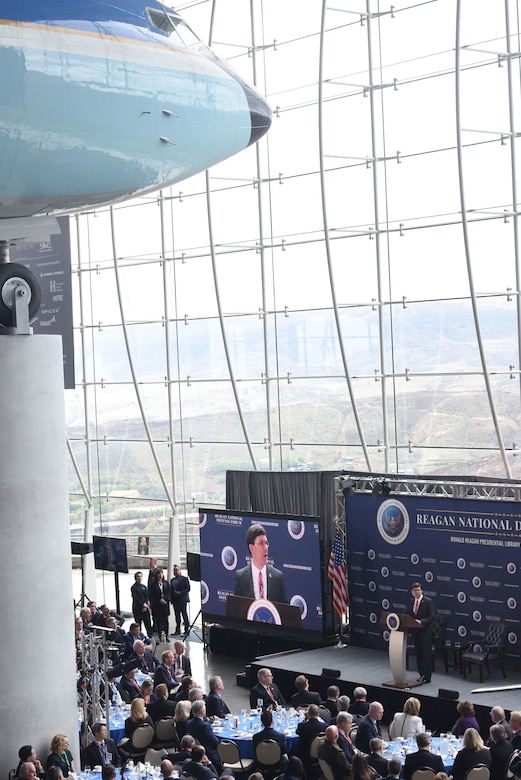 A man speaks at a podium while also being shown on a large screen to his right.