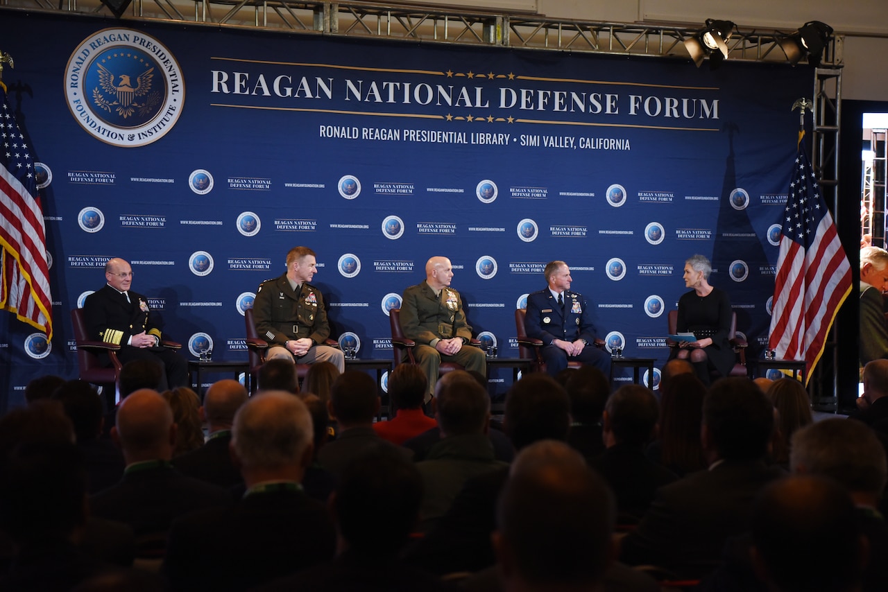 Men in military dress uniforms sit on a stage.