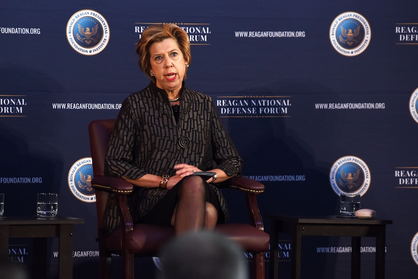 A woman sits in a leather chair. Behind her, a backdrop reads “Reagan National Defense.”