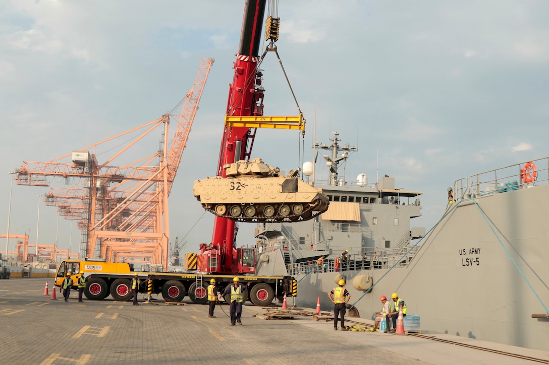 A crane lifts a tracked vehicle at a port as soldiers watch.