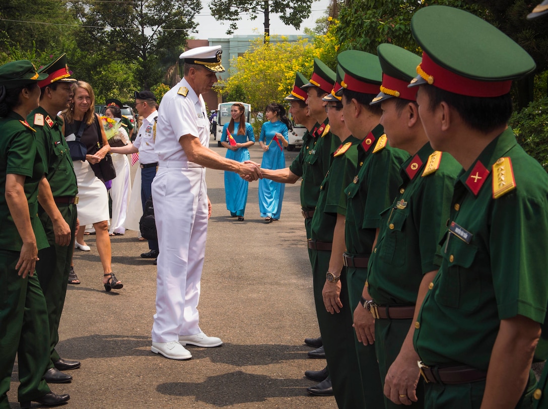 Navy admiral in white uniform shakes hands with one of several Vietnamese military personal lined up wearing green uniforms.