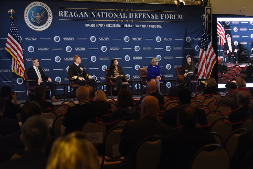 Navy admiral in uniform sits among four civilian panelists in front of an audience.