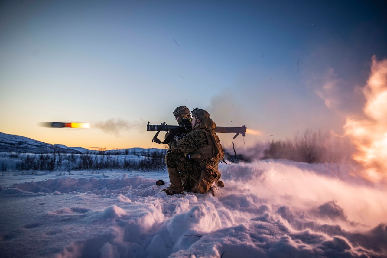 Two Marines kneel on a snowbank as a projectile emerges from a shoulder-launched weapon one of the Marines is firing.