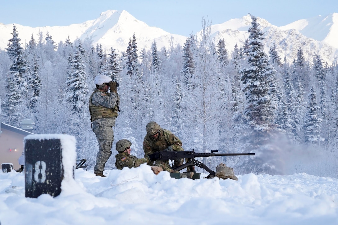 A soldier looks through binoculars as two soldiers fire a machine gun.