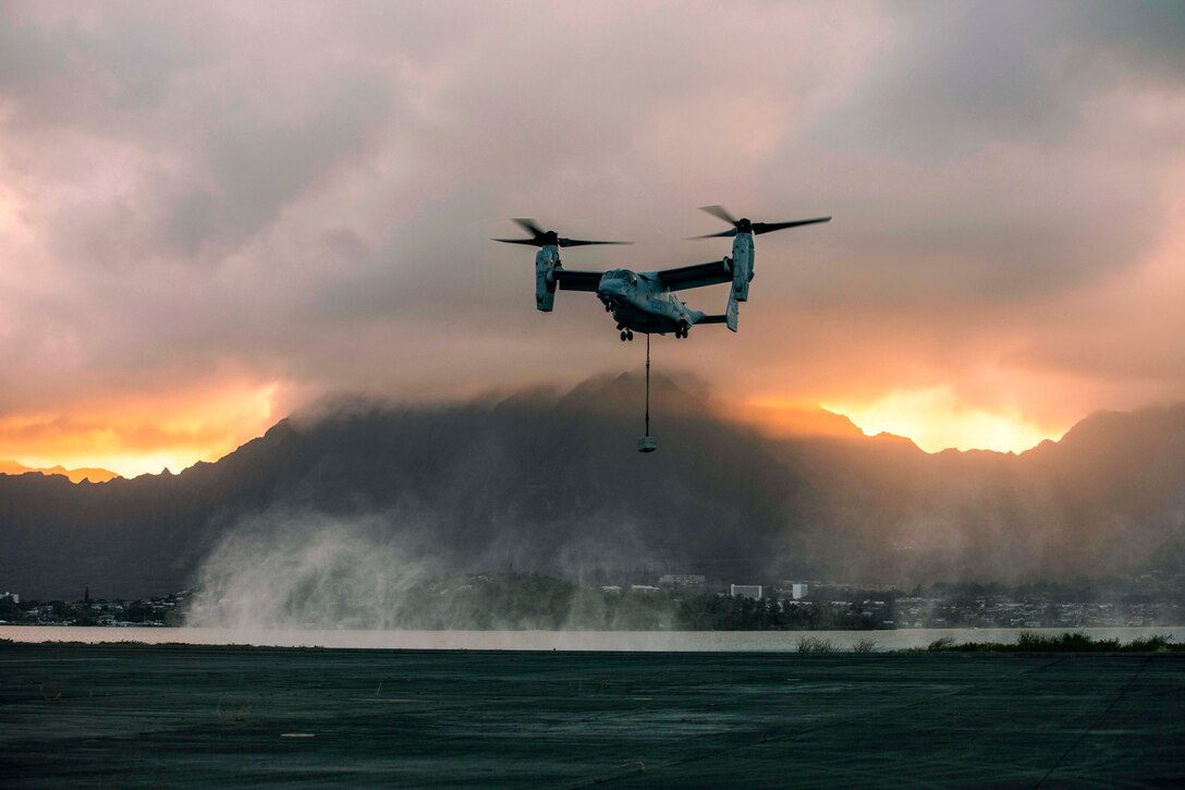 An aircraft carries a cement block hanging on a rope as the sun set.