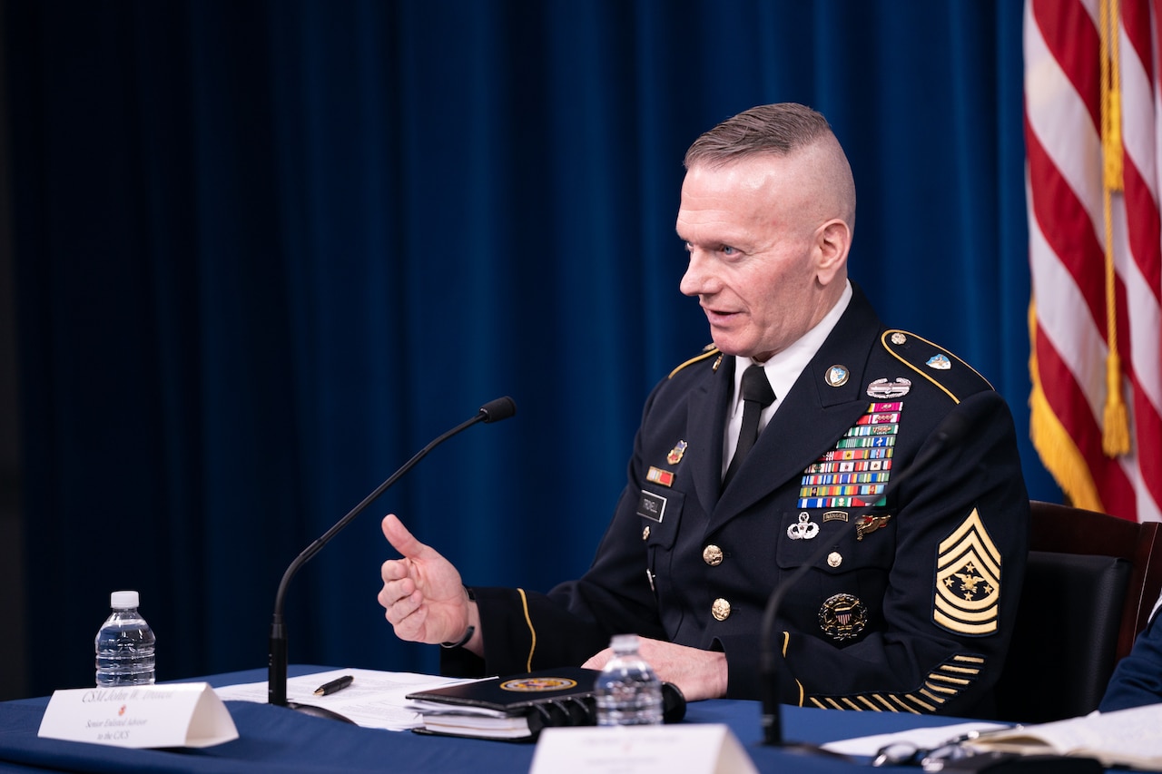 A uniformed service member sits behind a table and speaks into a microphone.