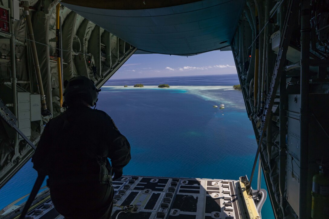 An airman looks out the back of a military plane as a parachuted bundle floats  through the air.