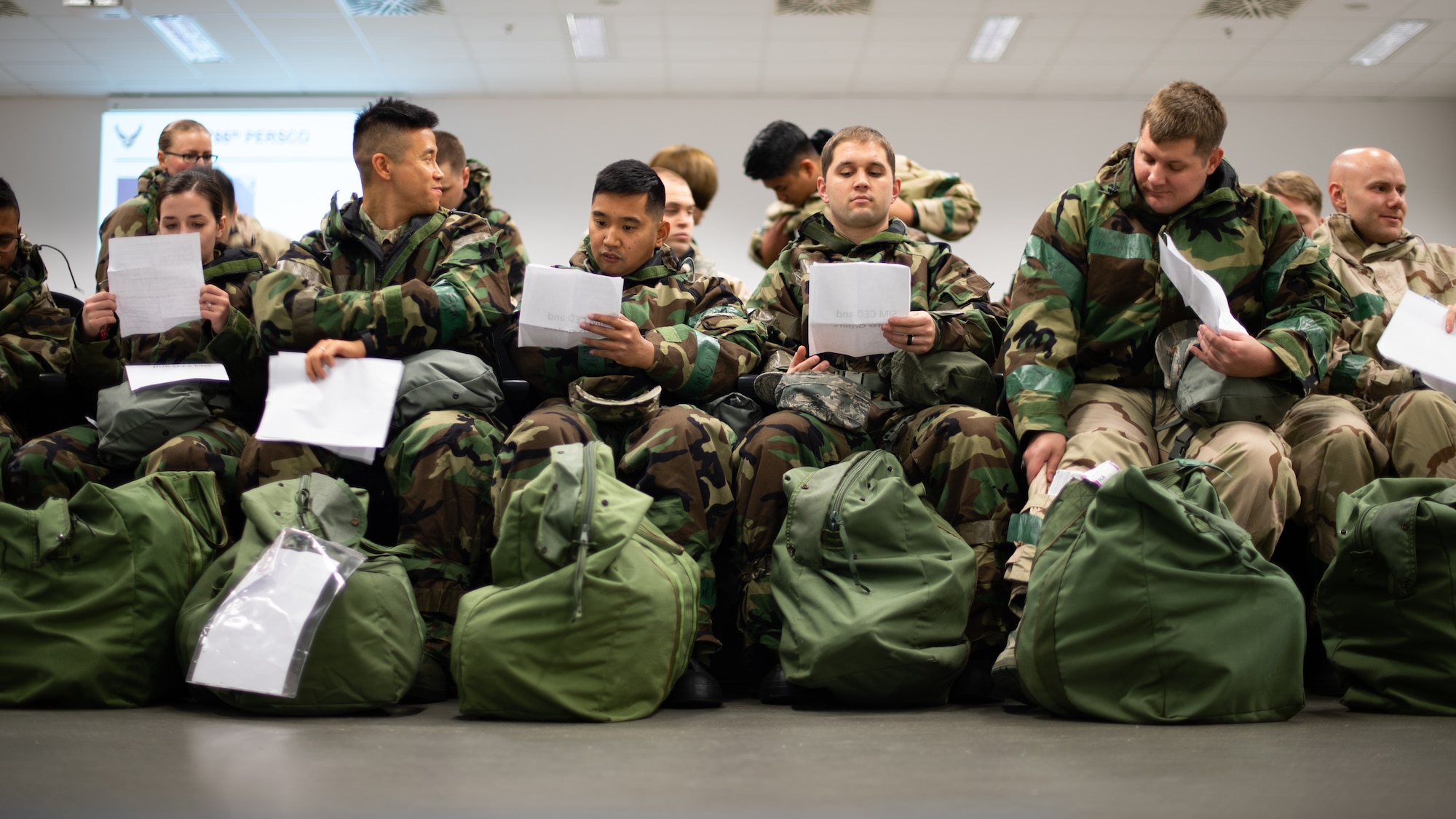 U.S. Air Force Airmen await a briefing at a simulated in-processing line during exercise Operation Varsity 19-04 at Ramstein Air Base, Germany, Dec. 9, 2019. Operation Varsity is a reoccurring exercise designed to enhance base response to a variety of attacks and situations including chemical, biological, radiological, nuclear, threats as well as the use of explosives. (U.S. Air Force photo by Staff Sgt. Devin Boyer)