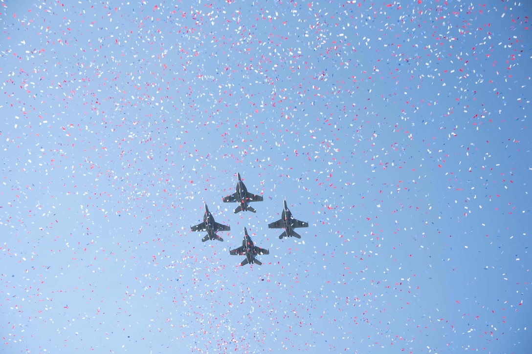 Four military jets fly through confetti against a blue sky.