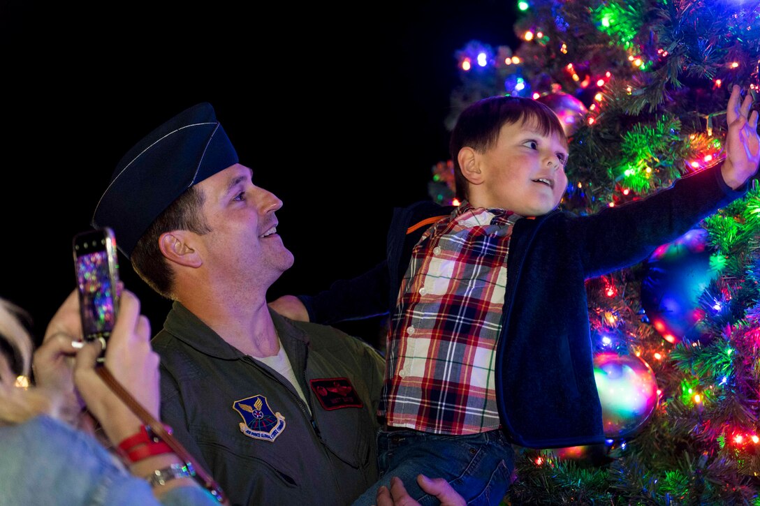 An airman holds a child next to a Christmas tree.