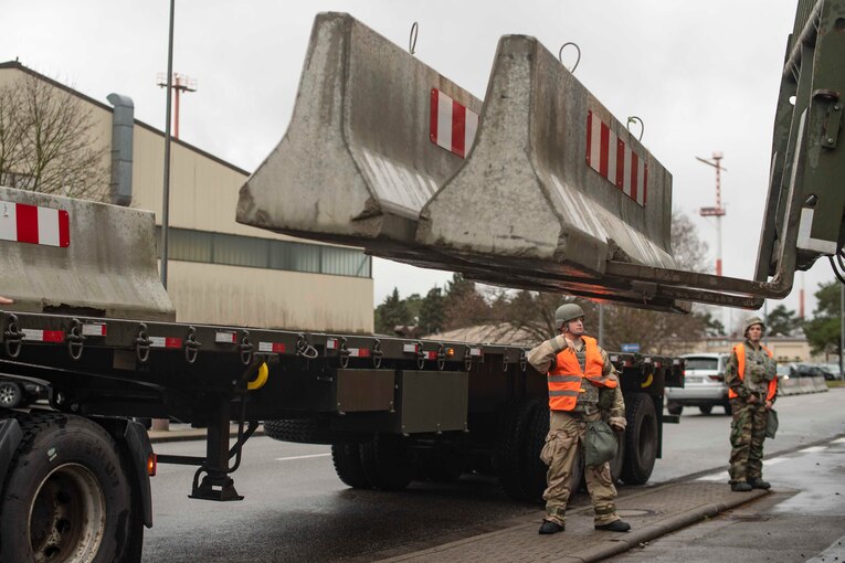 An airman stands next to a forklift as it moves concrete barriers.