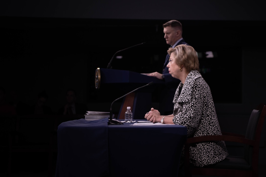 A woman sits at a table, with her hand folded.  In the background, a man in a military uniform stands behind a lectern.