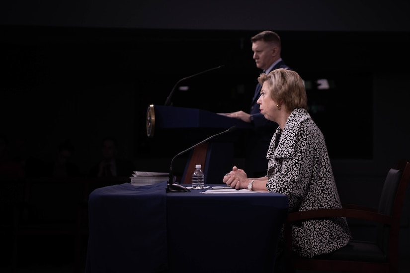 A woman sits at a table, with her hand folded.  In the background, a man in a military uniform stands behind a lectern.