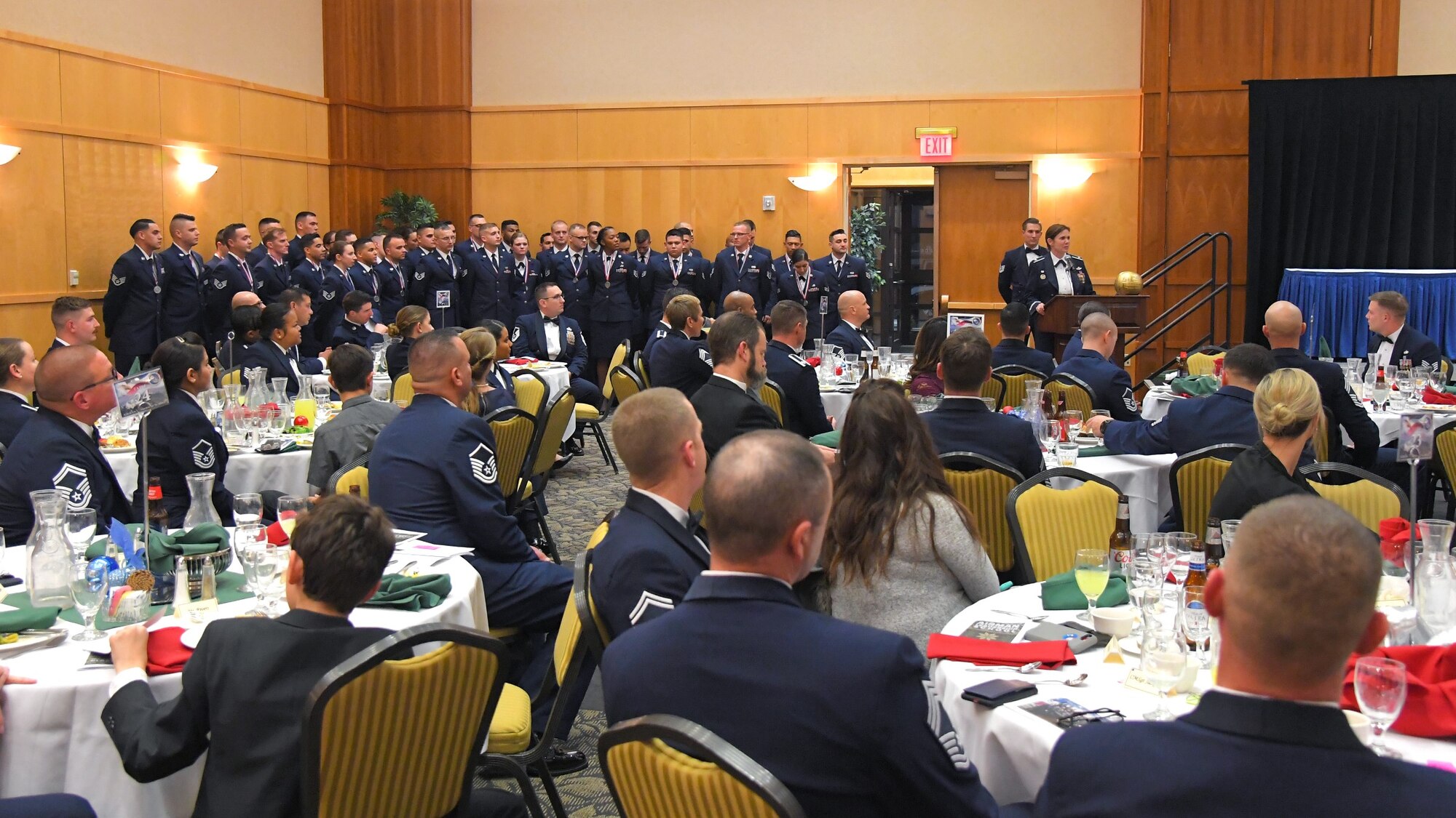 The graduates of Airman Leadership School Class 20-A pose for a group photo against ballroom wall while guests watch on from seated at a number of tables in the room.