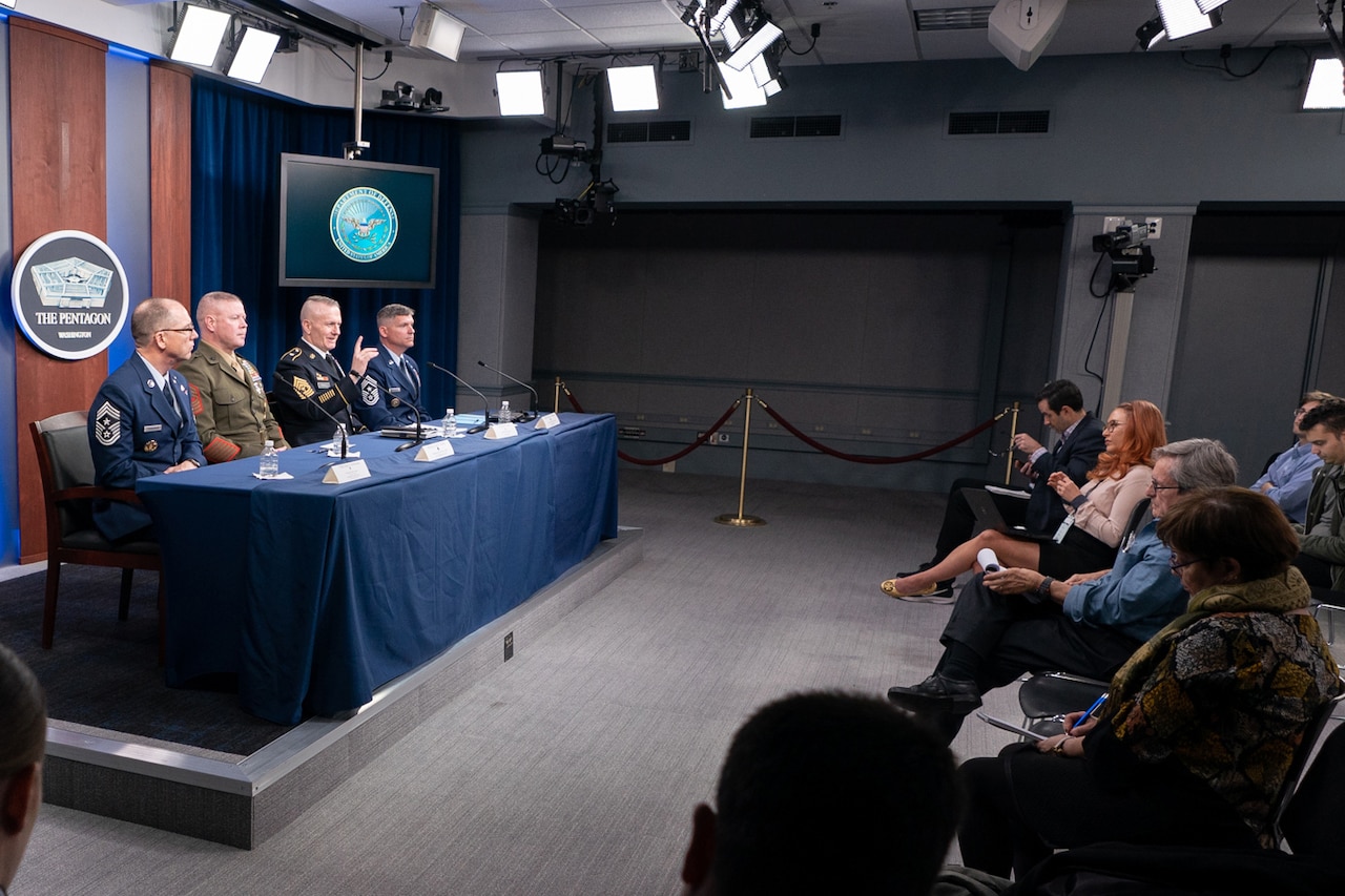 Four service members in uniform sit at a blue-draped table facing an audience of reporters.