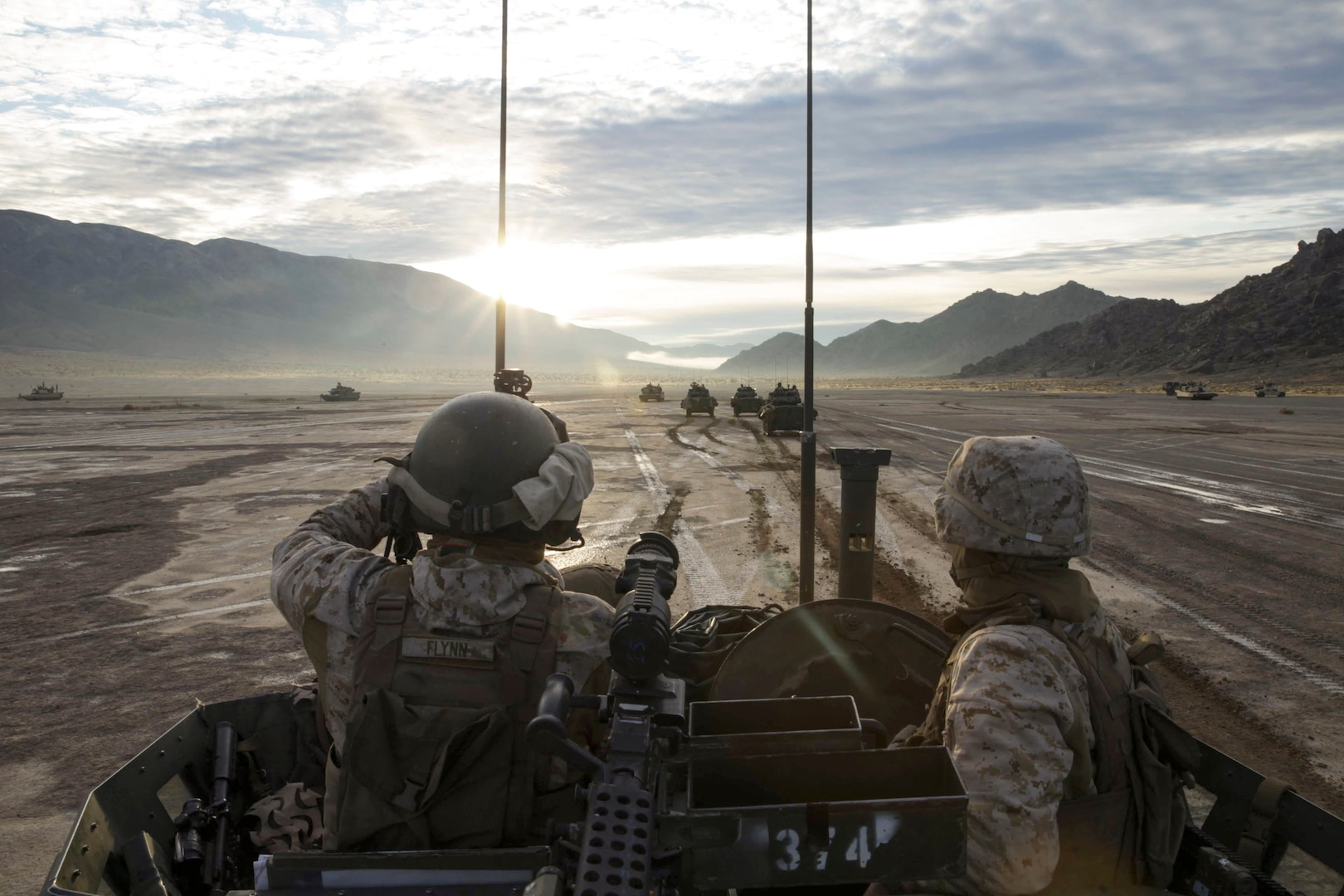 Two Marines ride in a tank in the desert as other tanks travel in the background.
