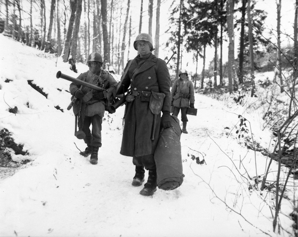 Three soldiers trudge through heavy snow in a forest carrying lots of equipment.