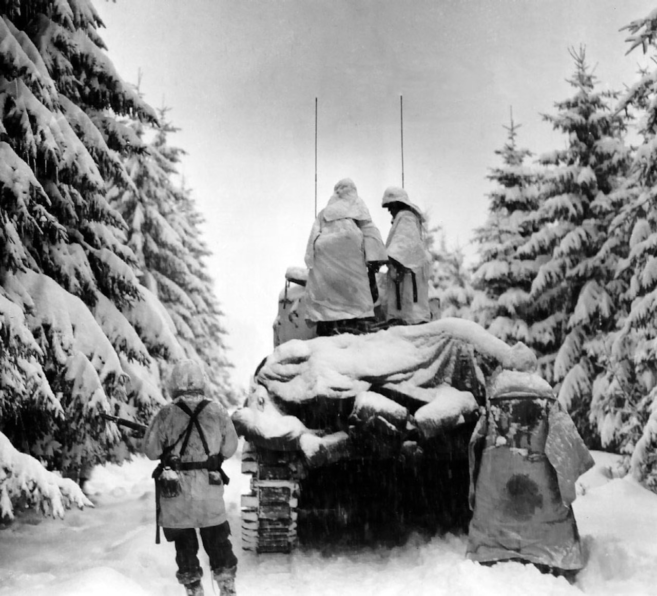 Two soldiers in winter clothing stand on top of a tank that’s pushing through heavy snow in a forest. Two other soldiers stand on the ground behind the tank.