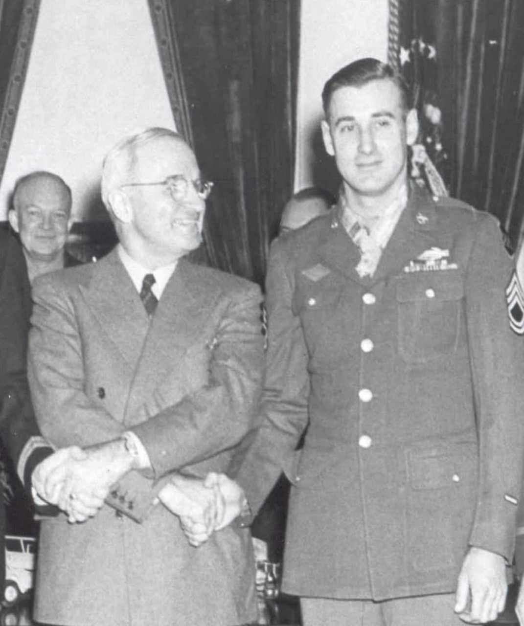 President Harry S. Truman, left, stands holding hands with and looking at a soldier to his left, who is wearing a Medal of Honor around his neck. Army Gen. Dwight D. Eisenhower is smiling in the background.