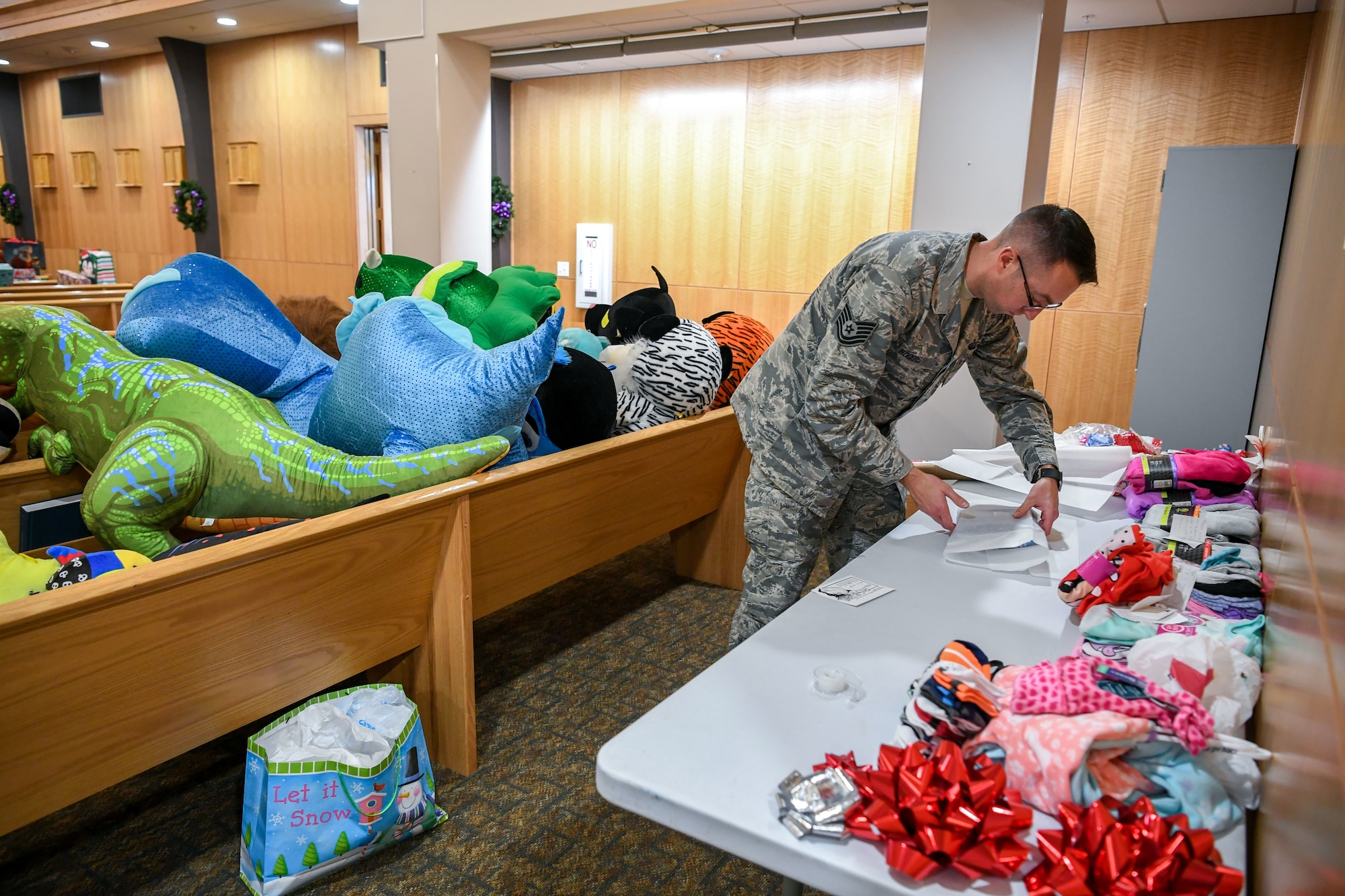 An Airman wrapping gifts on a table littered with cloths at the base chapel with stuffed animals filling pews behind him.
