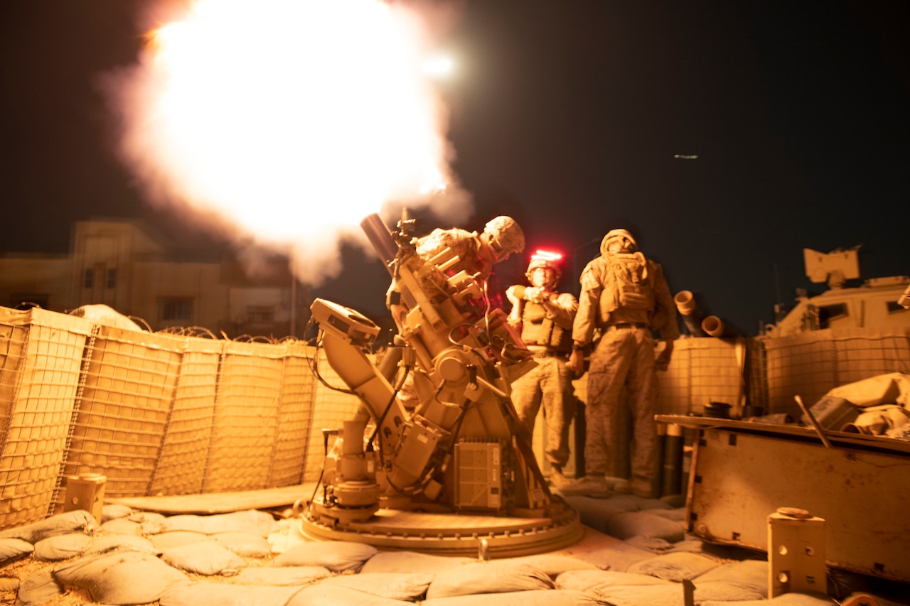 Service members fire a weapon at night. The explosion lights up the area.