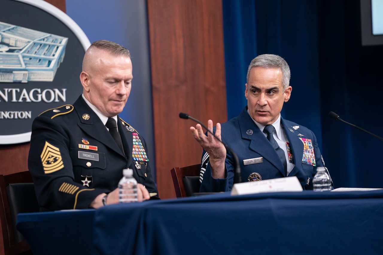 Two men in military uniforms sit at a table, behind microphones.