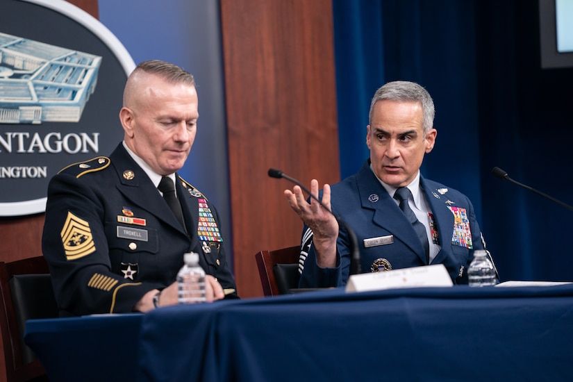 Two men in military uniforms sit at a table, behind microphones.