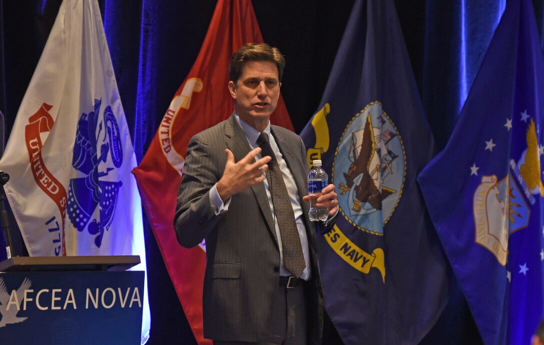 Man standing in front of four military service flags gestures while speaking to an audience