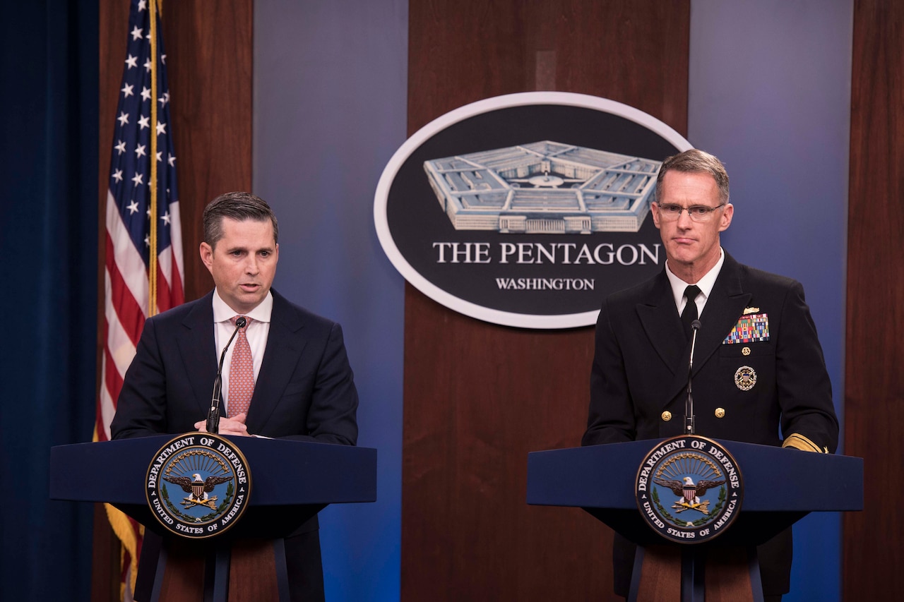 Two men, one wearing a military uniform, stand behind podiums and speak to reporters.