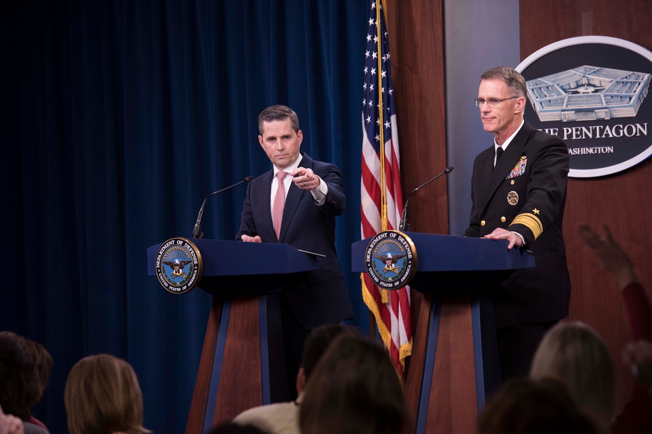 Two men, one wearing a military uniform, stand behind podiums and speak to reporters.