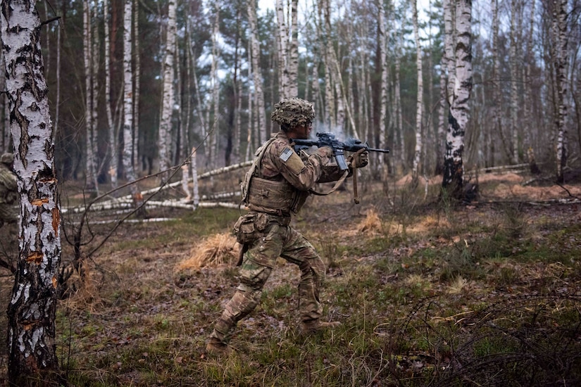 A soldier holds up a weapon while walking through a wooded area.