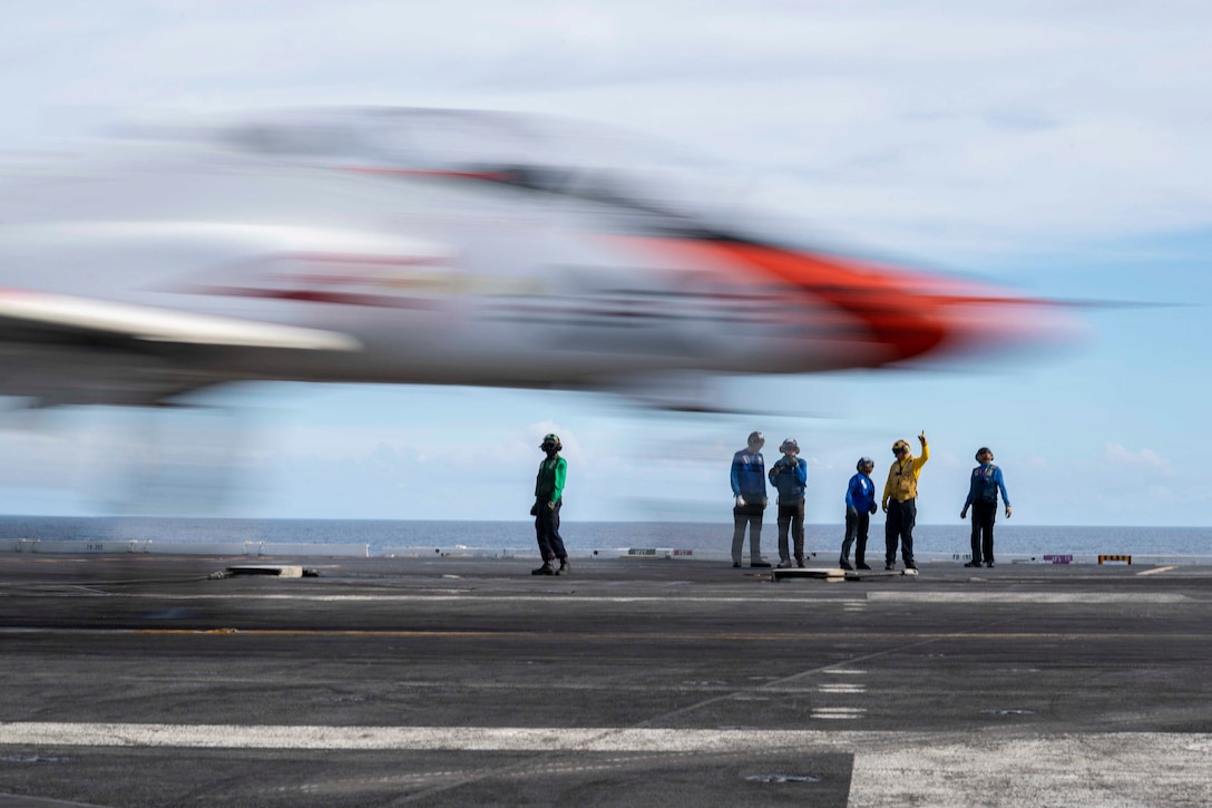 Sailors stand on a ship watch a plane land.