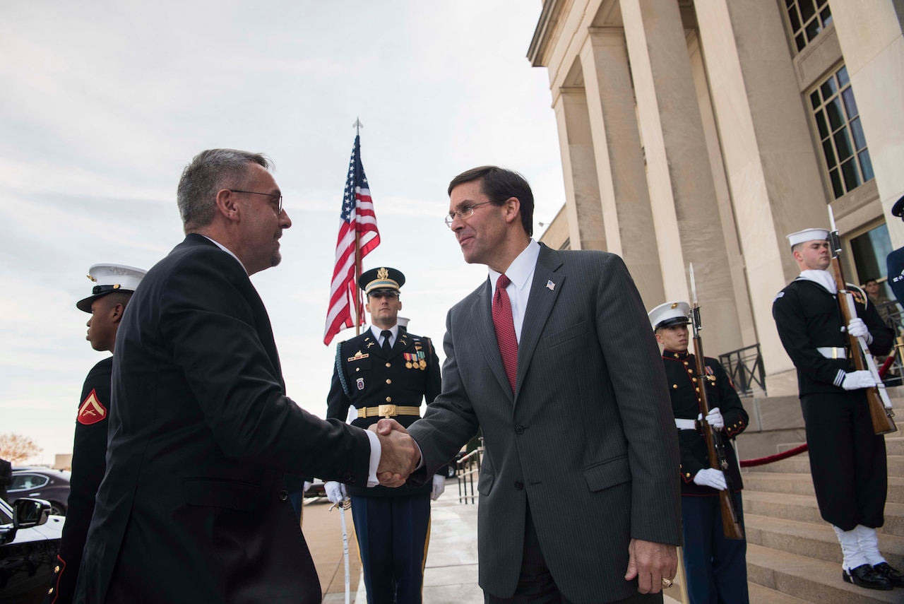 Two men shake hands. Military service members in dress uniforms stand in a row in the background.