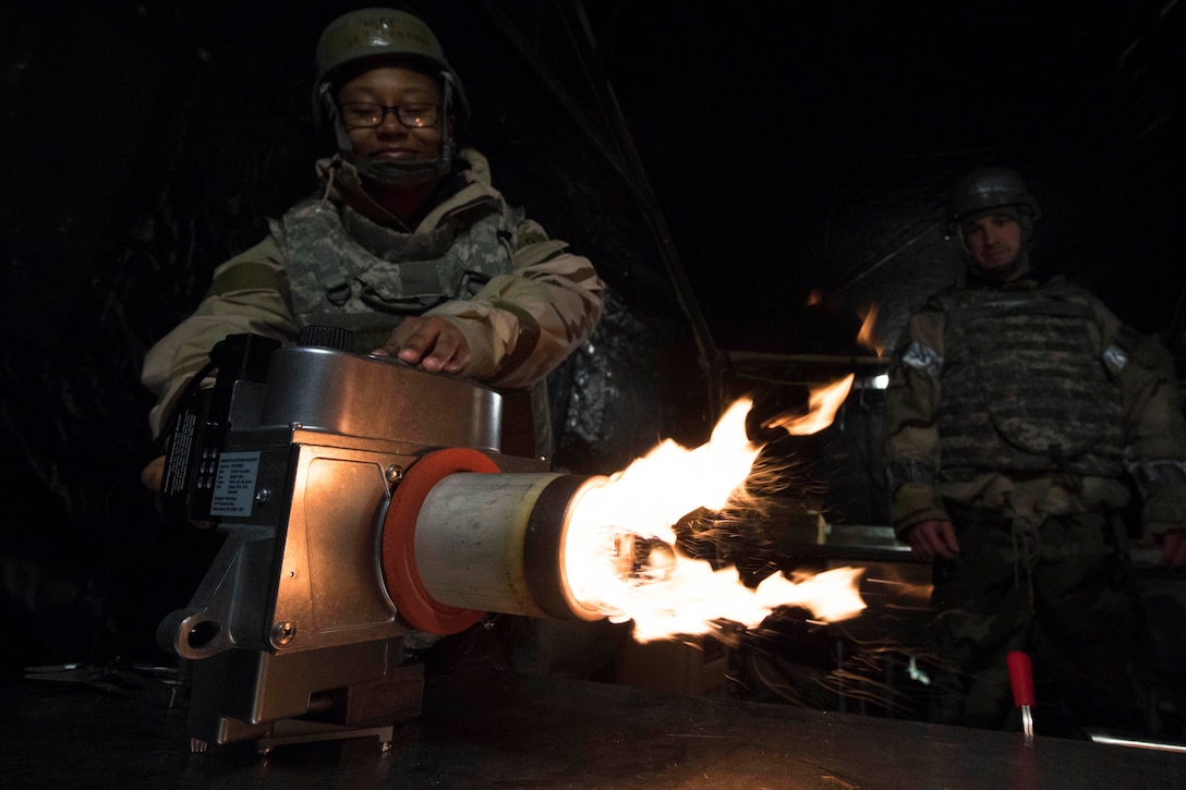An airman looks on while another lights a heater with flames shooting out.