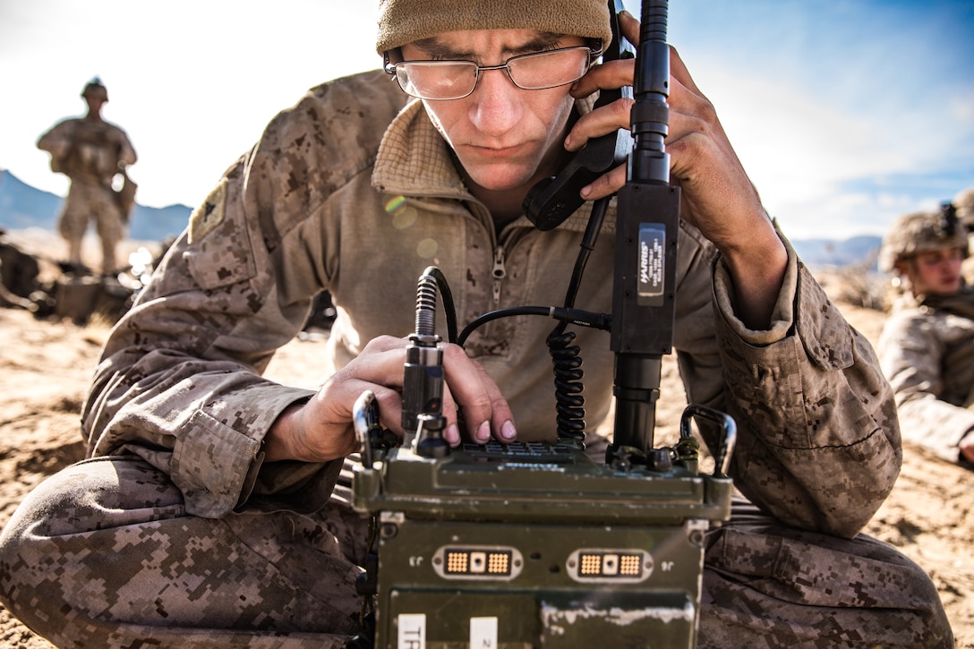 A Marine seated on the ground, works on a radio.