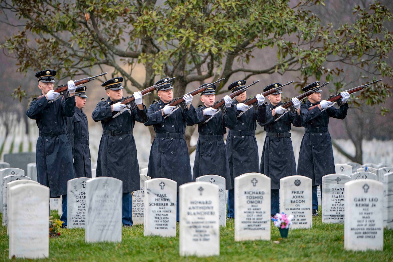 A group of soldiers stand in a line holding their rifles near tombstones.