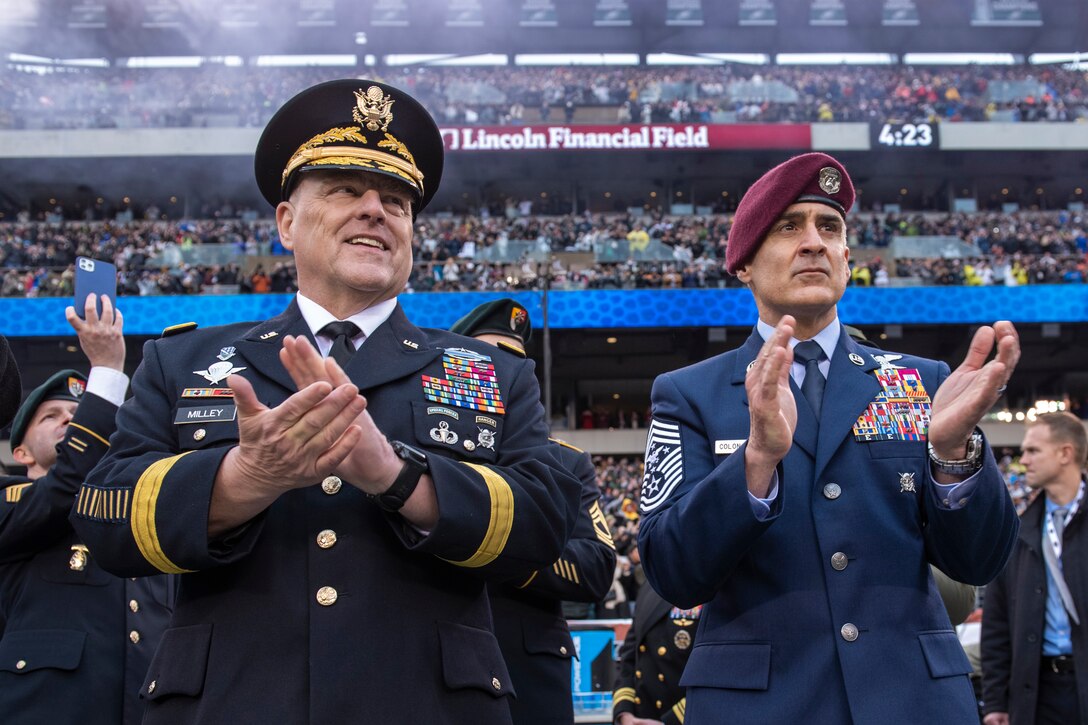 Service members in uniform clap while watching a football game, a crowded stadium in the background.