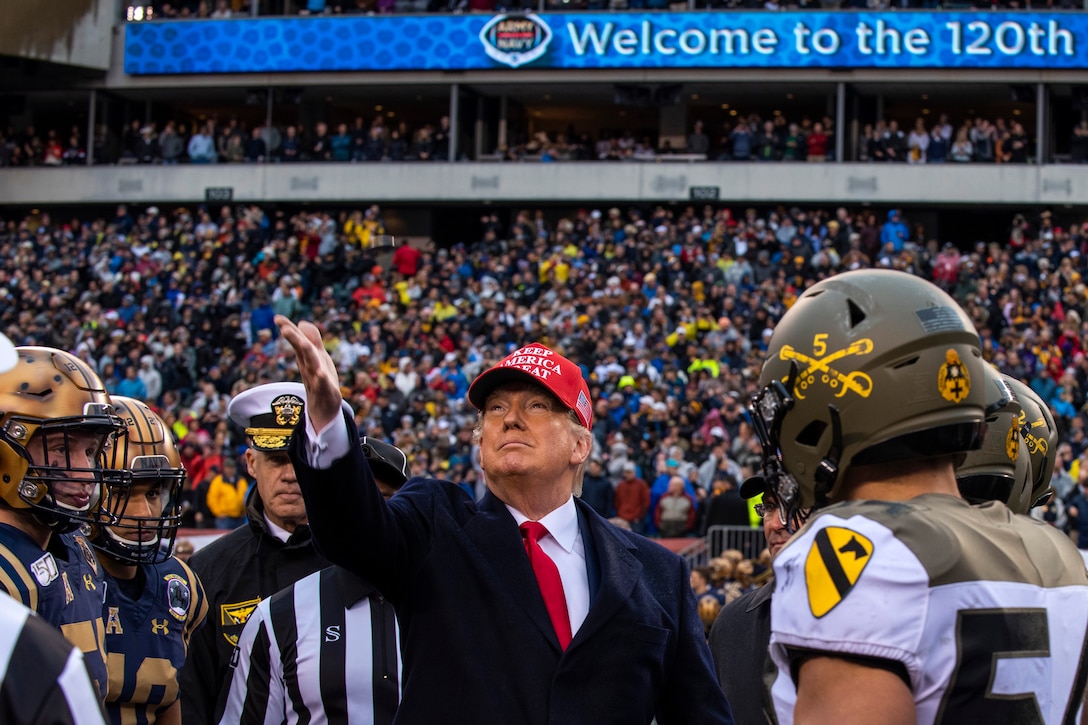 President Donald J. Trump, surrounded by people in football uniforms and a referee, tosses a coin.
