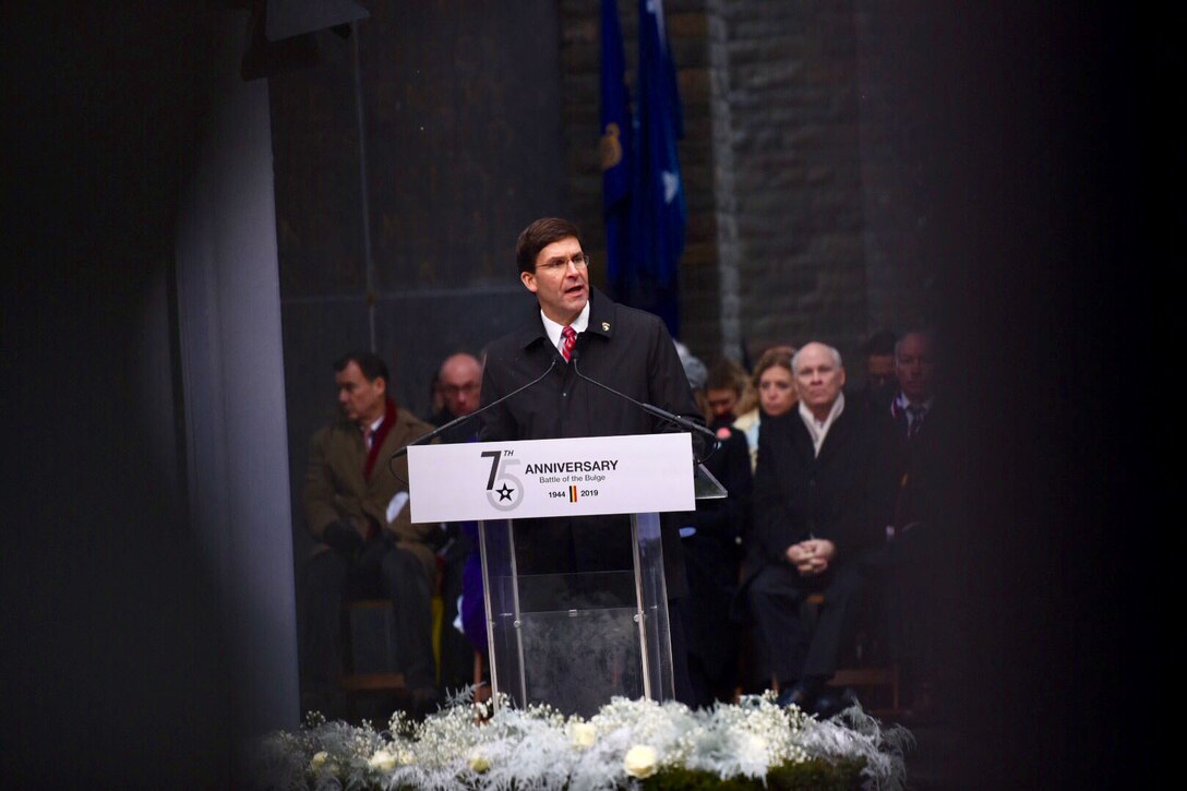 A man speaks from behind a clear podium. A sign on the podium reads, “75th Anniversary, Battle of the Bulge, 1944-2019.”