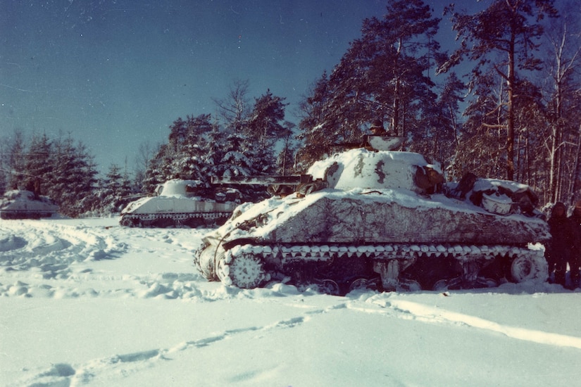 Several tanks sit in the snow in front of a line of trees.