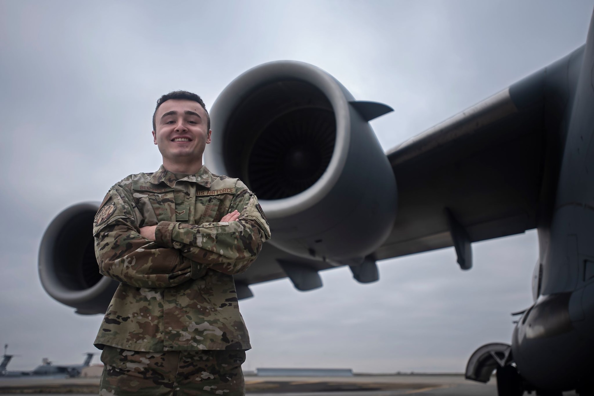 A man in a military uniform stands in front an aircraft's wing engines. It's overcast.