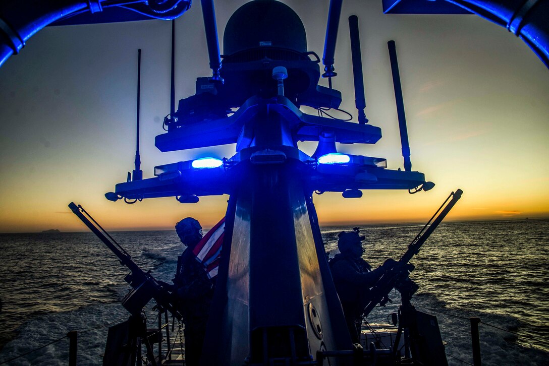 Two sailors stand on a ship at sea while the sun sets.