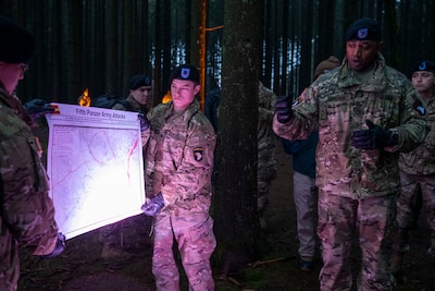 Soldiers hold a briefing chart titled “Fifth Panzer Army Attacks” as another soldier briefs visitors at a World War II battle site.