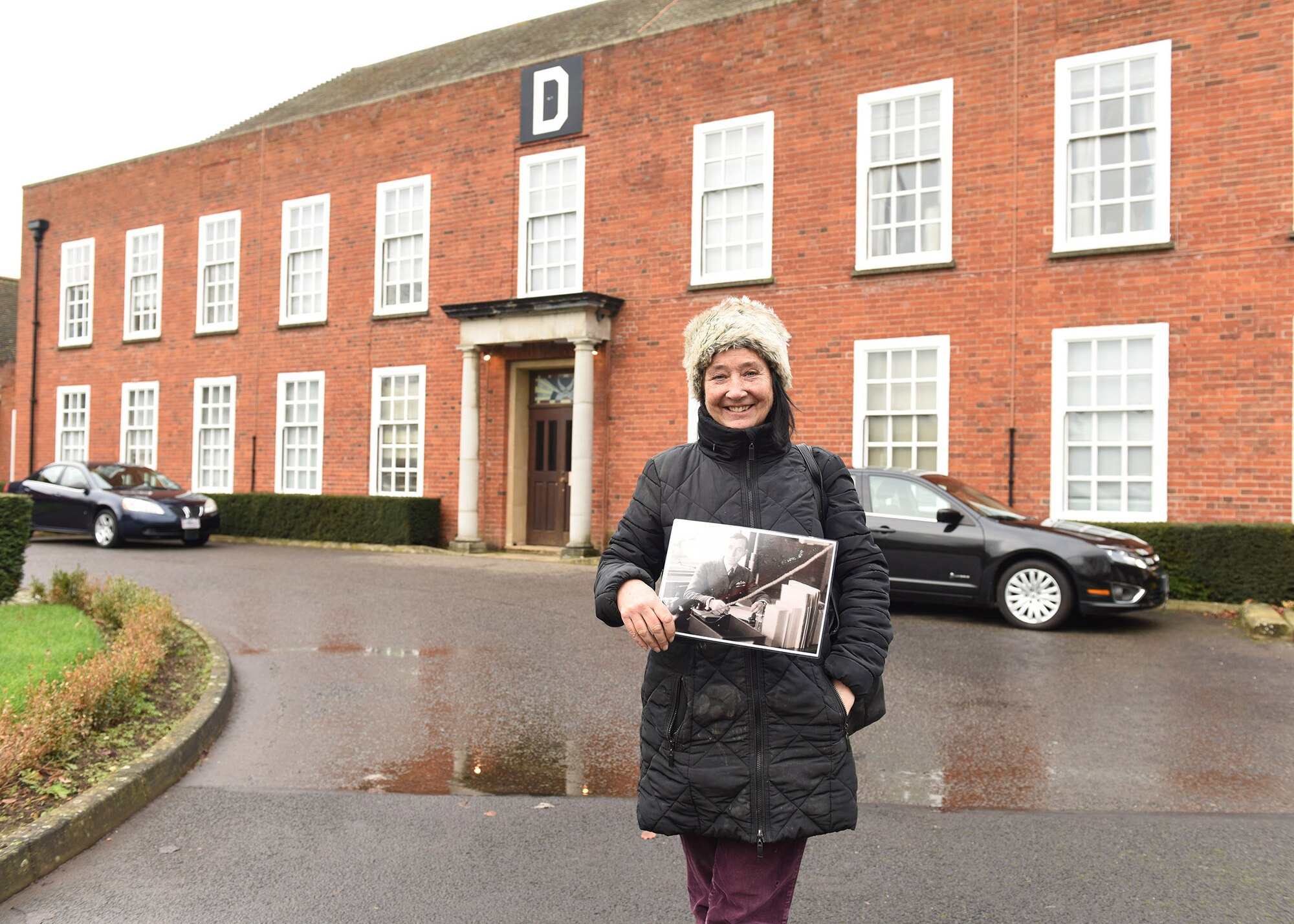 Rachel Kellett, niece of RAF Wing Commander Richard Kellett, poses with a photo of her uncle from his service during World War II, which shows him working in the 100th Air Refueling Wing headquarters building, then RAF Bomber Command headquarters, during a visit for members of The Heligoland 39 Project Dec. 16, 2019. Wing Commander Kellett flew the lead aircraft in daylight bombing raids on German ships during the first named battle of World War II, the Battle of Heligoland Bight. (U.S. Air Force photo by Karen Abeyasekere)