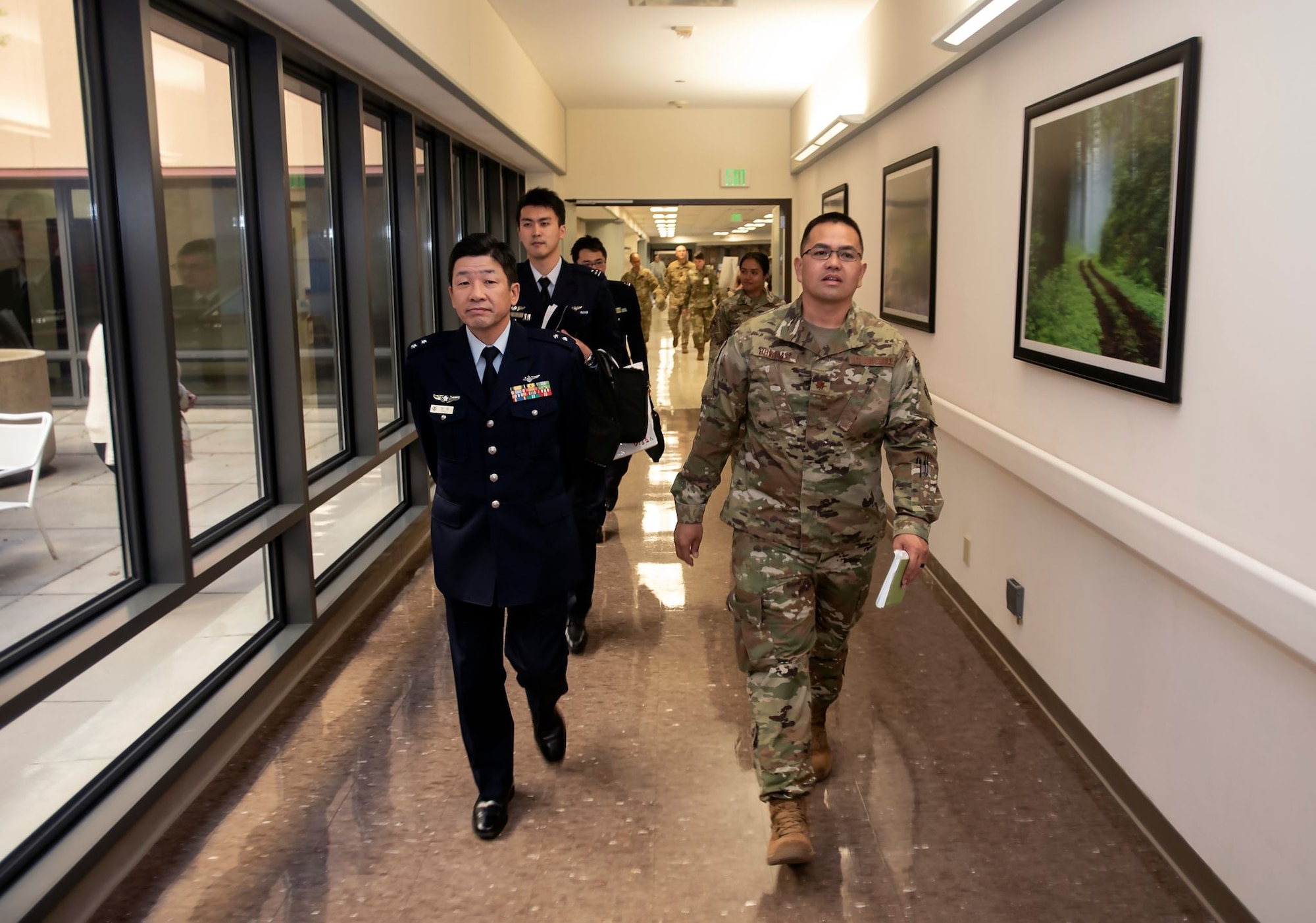 Two military personnel, one in dress blues and one in combat dress, walk down a long, brightly-lit hallway followed by a gaggle of other personnel