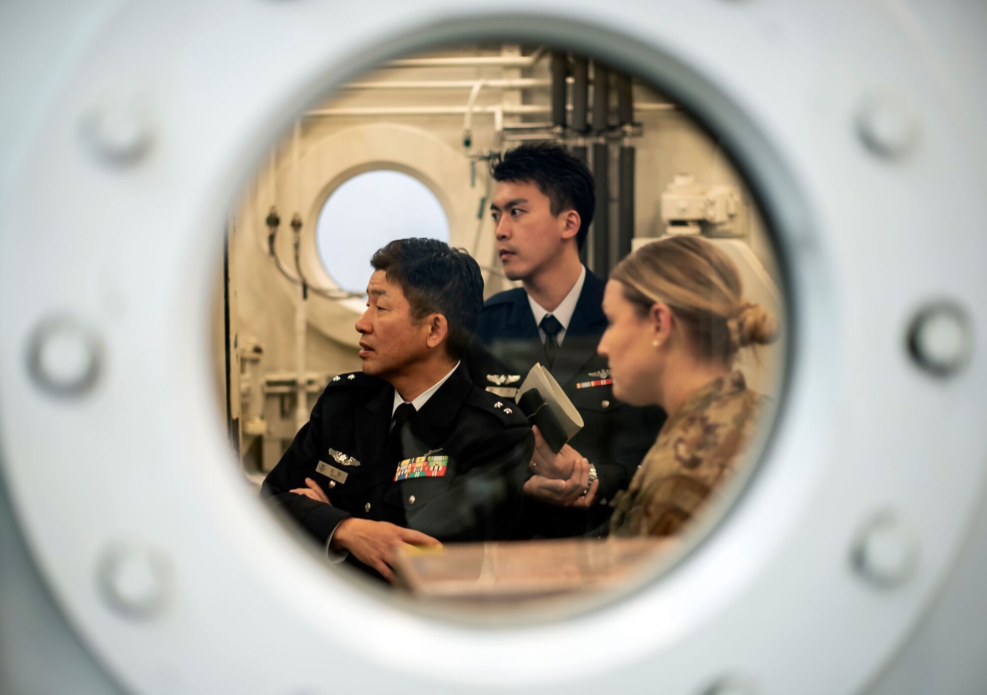 Three military personnel, two in dress uniform and one in combat uniform, are inside a pressurized tank apparatus